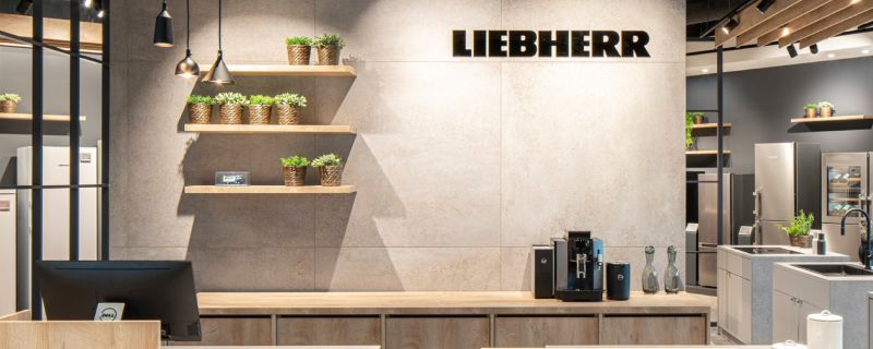 Empfangsbereich mit LIEBHERR-Logo, Pflanzen auf Regalen, Kaffeemaschine auf Theke. Empfangsbereich mit LIEBHERR-Logo, Pflanzen auf Regalen, Kaffeemaschine auf Theke.
